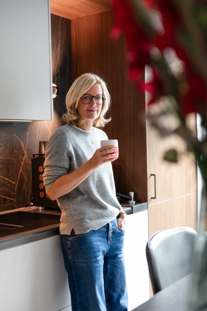 Corinna Starcke steht mit einer Kaffeetasse in der Küche, rote Blumen im Vordergrund